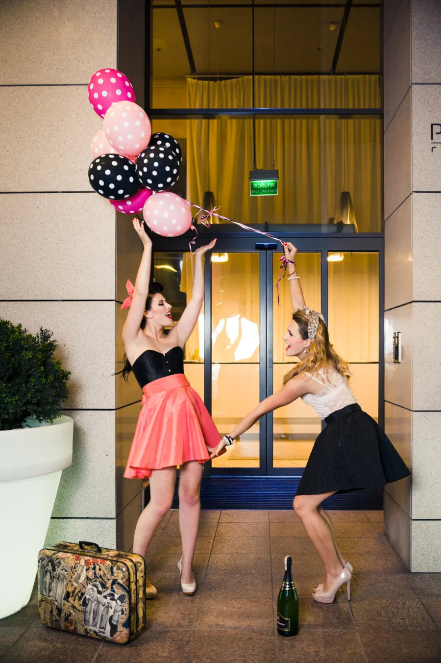 Two women dressed stylishly in party attire stand in front of a building. One is holding a bunch of colorful balloons, the other is reaching out to catch them. Next to them on the ground lies an antique suitcase and a bottle of champagne, perfectly captured by well-known Warsaw photographer Marcin Krokowski.  