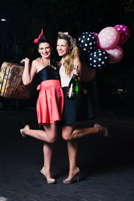 Two stylishly dressed women smile and pose outdoors at night. One holds a vintage suitcase, the other carries balloons and a large bottle of champagne. Both have a playful leg up, creating a joyful and celebratory atmosphere that was perfectly captured by Marcin Krokowski, a well-known photographer in Warsaw.  