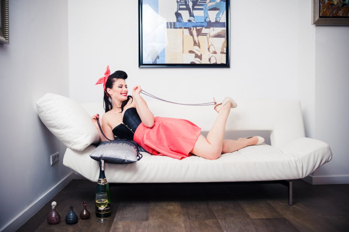 A woman with a retro hairstyle and bright red lipstick lies on a white sofa and playfully holds a string of pearls. She is wearing a black strapless top and a red skirt with one leg raised playfully. This charming moment was captured by Marcin Krokowski, a well-known photographer in Warsaw. Behind her, a painting hangs on the wall and bottles stand on the floor.   