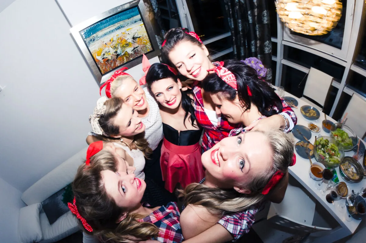 A group of seven smiling women, dressed in vintage-style clothing and red headbands, gather closely in a well-lit room. Near a table set with plates of food, one woman is wearing a distinctive black and pink dress. This charming moment was captured by Marcin Krokowski, a well-known photographer in Warsaw.  