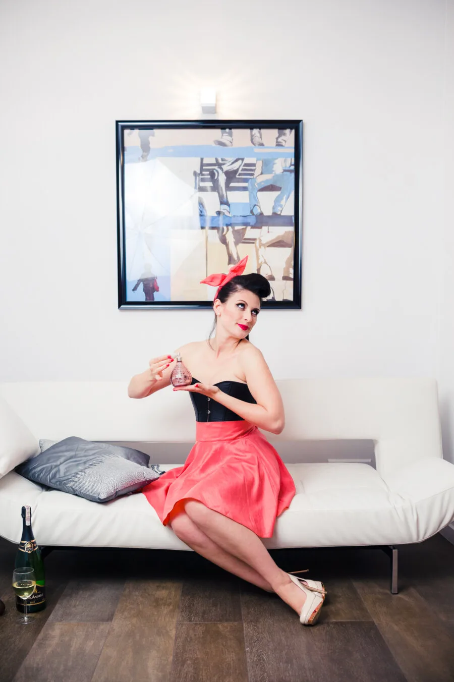 A woman with vintage makeup and hairstyle sits on a white couch. She holds a bottle of perfume in her right hand, wearing a black top, red skirt and red bow. On the floor lies a bottle of champagne and two glasses. Behind her on the wall hangs a contemporary print, brilliantly captured by Marcin Krokowski.   