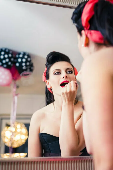 A woman with a red headband applies red lipstick while looking in the mirror. She is wearing a black strapless top and her dark hair is pulled back. In the background are fuzzy balloons and lighting, skillfully captured by photographer in Warsaw Marcin Krokowski.  