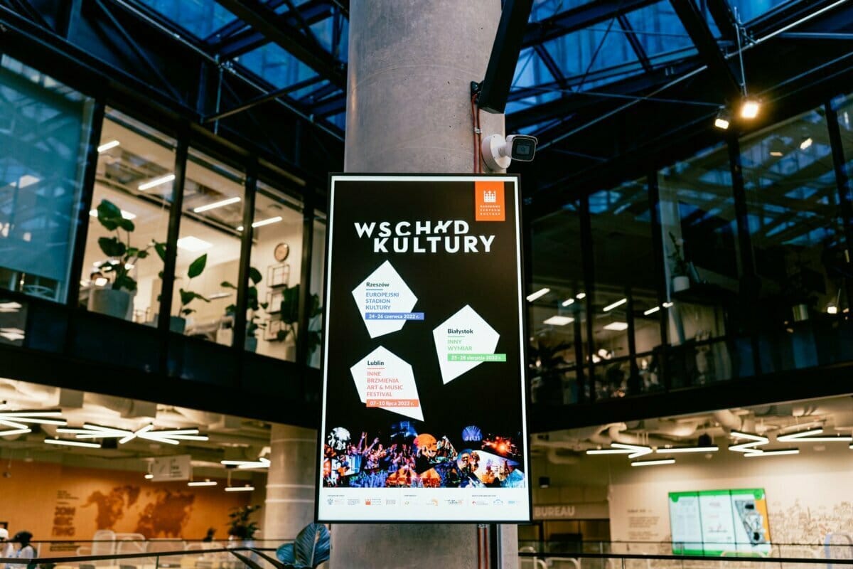 The poster is displayed in a modern glass building. Entitled "EAST OF CULTURE," the poster features various geometric shapes with city names and event details in Polish, and at the bottom images of the performing band and cultural icons, captured by well-known event photographer Marcin Krokowski. 