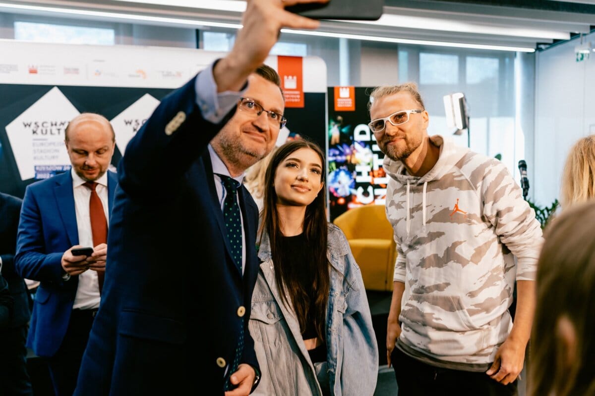 A group of three people stand together, posing for a selfie. A man in a suit is holding a phone, a young woman in a denim jacket stands in the middle, and a man in a white and gray moro sweatshirt stands to the right. Other people and a modern background are visible, beautifully captured by event photographer Marcin Krokowski.  