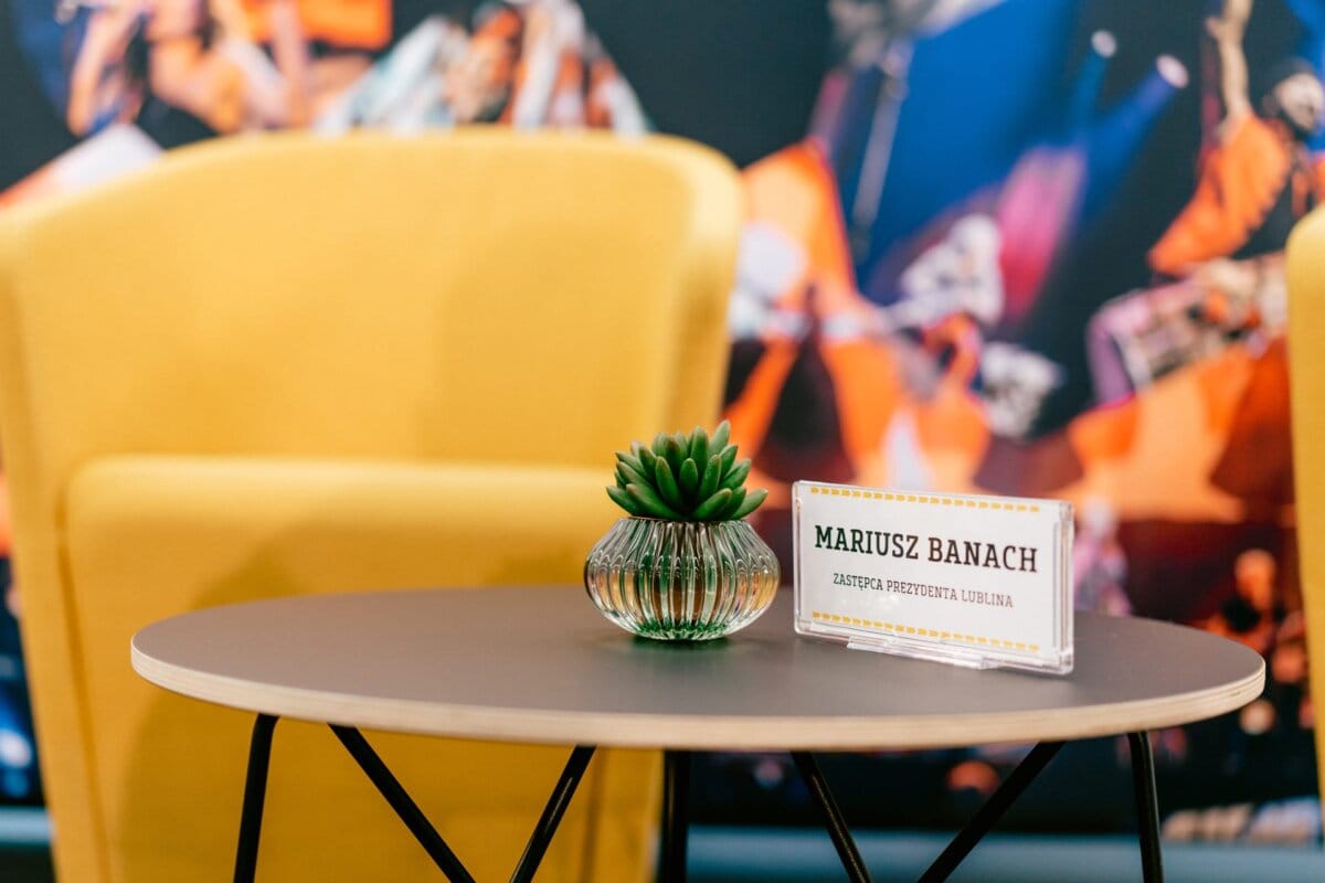 A small round table with a succulent in a pot in a glass vase and a plaque with the inscription "Mariusz Banach" placed on it. On either side of the table are two yellow chairs, prepared for an elegant meeting arranged by Marcin Krokowski, a photographer in Warsaw. A colorful, fuzzy background is visible.  