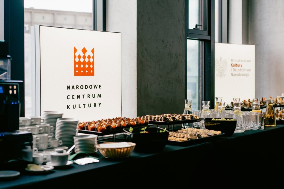 The table is set with various foods and drinks, including cakes, small snacks and beverages. Behind the table are two illuminated signs with the logos and names of the "National Cultural Center" and the "Ministry of Culture and National Heritage," which were brilliantly captured by Marcin Krokowski, an acclaimed Warsaw event photographer. 