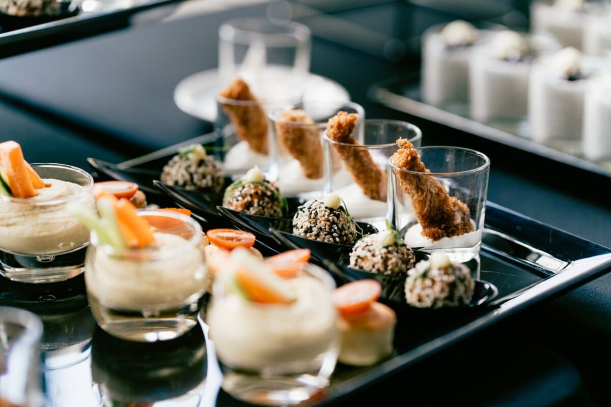 Elegant appetizers on the buffet table, consisting of hummus with chopped vegetables in small dishes and clear glasses filled with something resembling a creamy dip with fried produce. In the background, other small appetizers, beautifully captured by photographer in Warsaw Marcin Krokowski. 