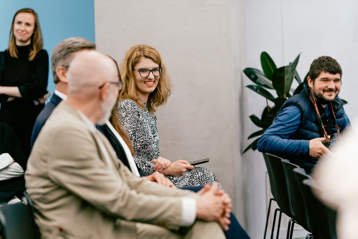 A group of people sitting in a room. A woman wearing glasses and a patterned dress smiles and interacts with others. Two men can be seen in the back, one in a beige and the other in a gray suit. Marcin Krokowski, a well-known Warsaw photographer, is holding a camera in a blue vest. Two women stand in the background.    