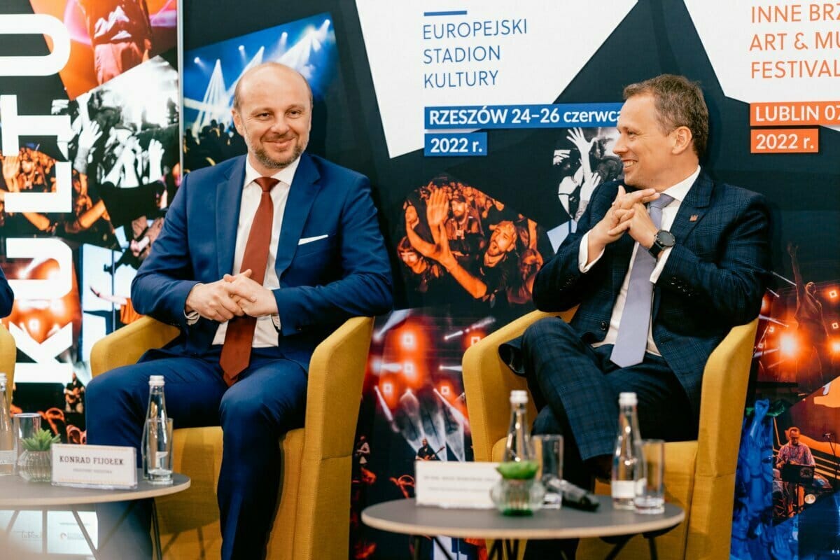 Two men in suits sit on yellow chairs, smiling and carrying on a conversation. Behind them, colorful banners advertise cultural and musical events, bearing the words "EUROPEAN STADIUM OF CULTURE" and dates for June 2022. This vivid moment is captured by Marcin Krokowski, a well-known Warsaw photographer.  