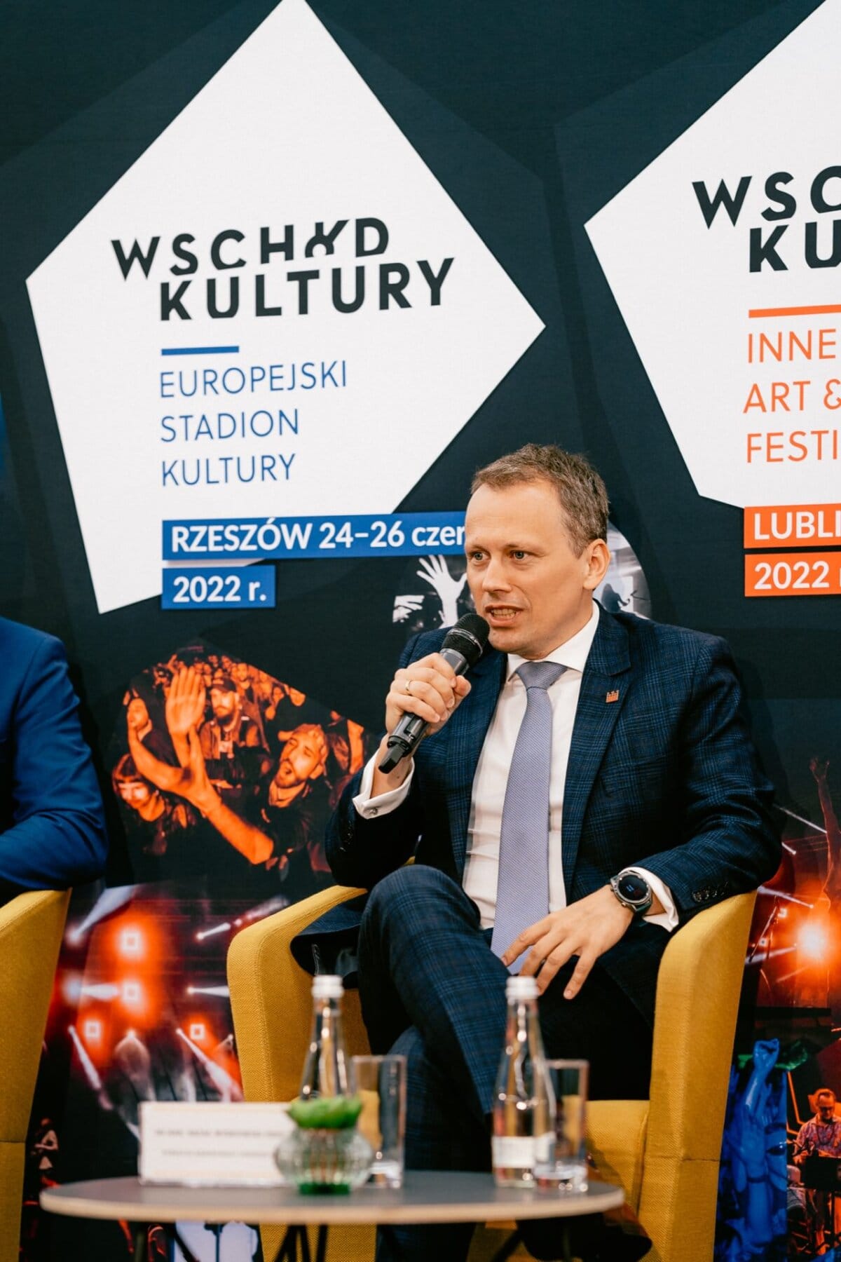 A man in a suit speaks into a microphone while sitting on a yellow chair at a cultural event. Behind him, two posters hang "East of Culture European Stadium of Culture Rzeszow, June 24-26, 2022" and "East of Culture Other Days Art &amp; Music Festival Lublin 2022." Marcin Krokowski, photographer in Warsaw  