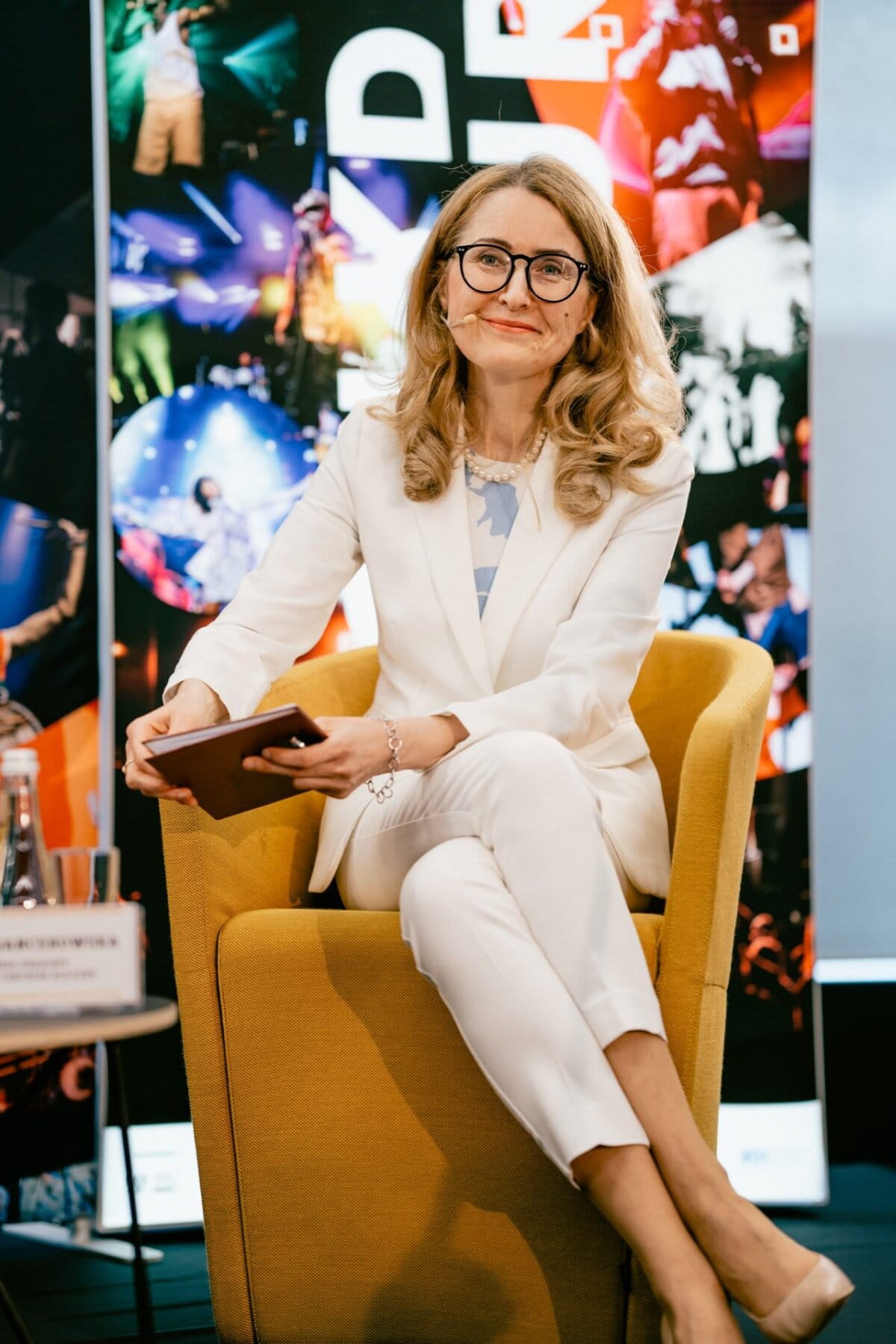 A woman with long blond hair and glasses sits in a yellow chair and holds a small notebook. She is wearing a white suit and smiling. In the background is a colorful collage of various photos, taken by well-known Warsaw event photographer Marcin Krokowski. The nameplate is partially visible on the table next to her.   