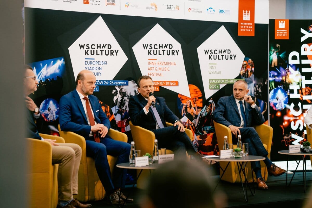 A panel of four men in suits take part in a discussion at a cultural festival. They are sitting on yellow chairs on the stage, behind them colorful banners with the words "EAST OF CULTURE" and the dates of the events. This moment was brilliantly captured by Marcin Krokowski, a well-known photographer in Warsaw.  