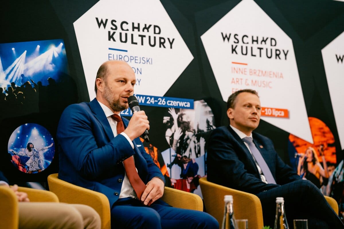 Two men in suits sit and speak during a panel discussion. Behind them, banners display information about "East of Culture" festivals, including "European Stadium of Culture" and "Other Sounds of Art &amp; Music" with dates "June 24-26" and "July 7-10." The event was captured by Marcin Krokowski,  