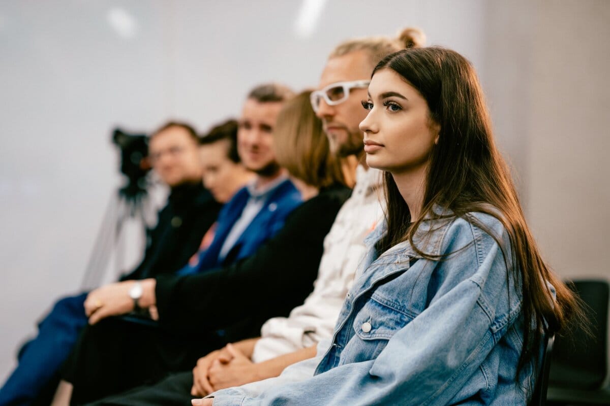 A group of people sit in a row and listen intently, while most look ahead. We focus on a young woman in the foreground, wearing a denim jacket. In the background, a camera set up on a tripod suggests that Marcin Krokowski, a well-known photographer in Warsaw, is covering the event.  