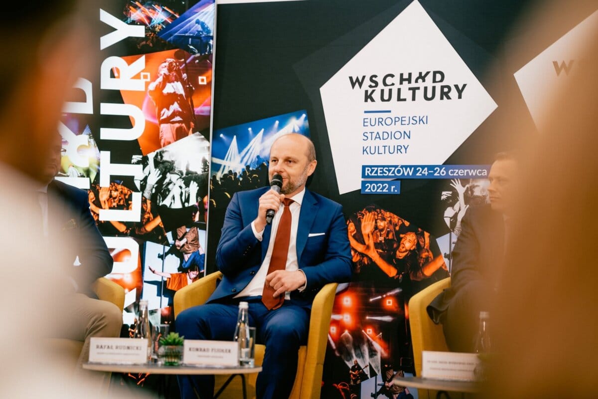 A man in a blue suit speaks into a microphone while seated during a panel event. In the background are vivid images and texts promoting the "East of Culture" festival, which will be held in Rzeszow from June 24-26, 2022. Marcin Krokowski, a well-known Warsaw-based event photographer, captures the moment among the other panelists.  