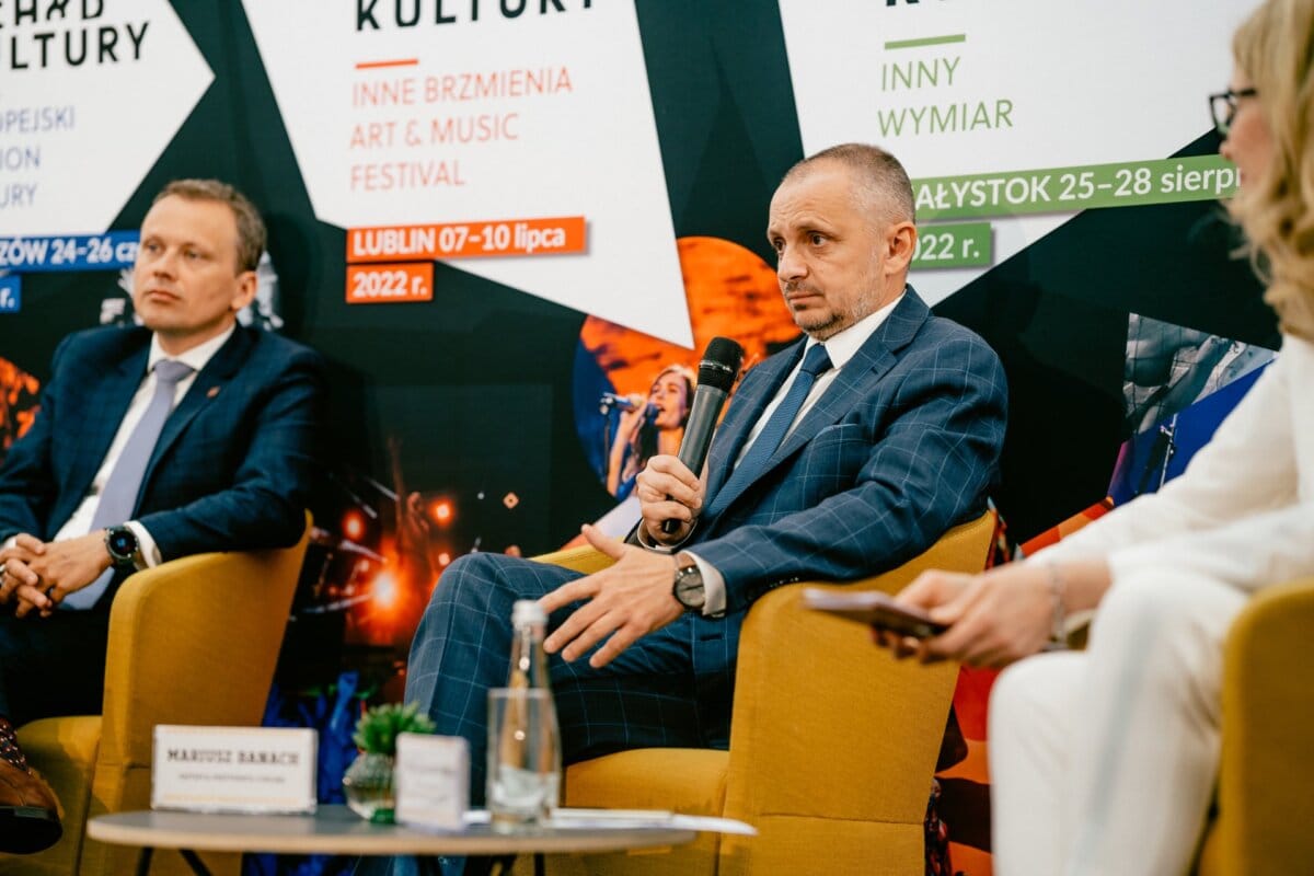 On stage, three people in formal attire sit in chairs. One man in a blue plaid suit, holding a microphone, appears to be speaking. Behind them hang posters of events with dates and locations, including "photographer in Warsaw" and Marcin Krokowski's name. On a table in front stand water bottles and a small plant.    