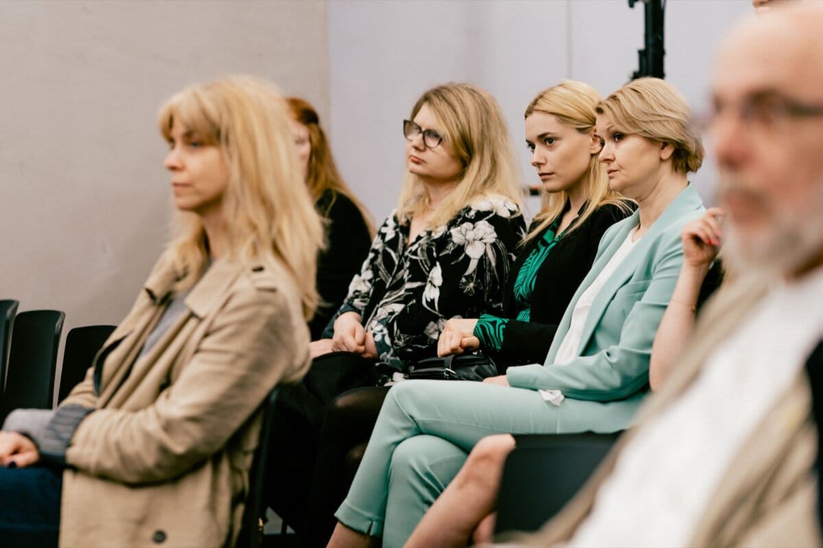 A diverse group of professionally dressed people sit attentively in a conference setting captured by acclaimed event photographer Marcin Krokowski. Some seem engaged and focused, while others look thoughtful. The background is neutral, with a light wall and equipment visible.  