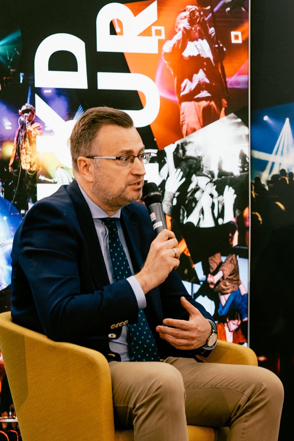 A man in a blue jacket, white shirt and green tie, holding a microphone, sitting on a yellow chair, speaks against a background filled with photos of concerts and musical performers captured by Marcin Krokowski, a well-known event photographer and photographer in Warsaw.