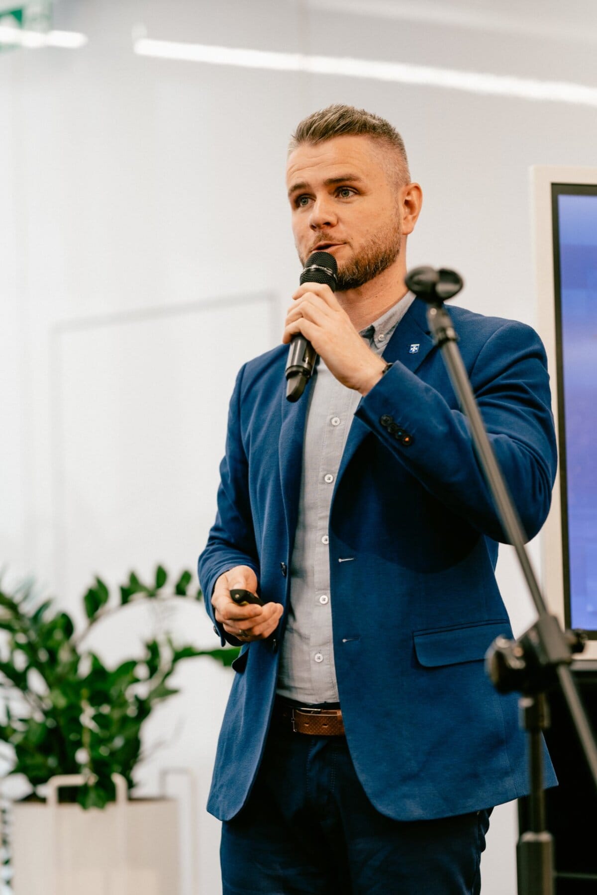 A man in a blue suit, probably Marcin Krokowski, is speaking into a microphone, holding a remote control in his other hand. He appears to be giving a presentation, with a plant and a large screen visible in the background. 