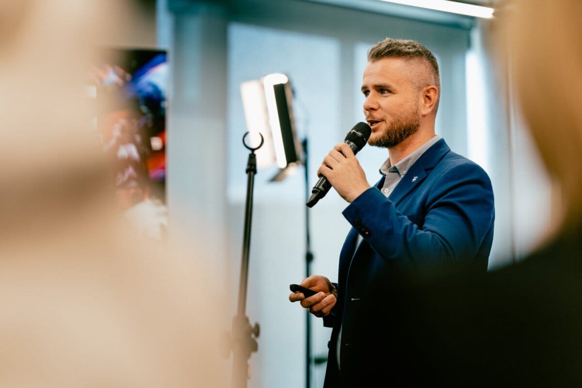 A man with a beard and short hair, wearing a blue jacket, speaks into a microphone while holding a remote control. He stands in a well-lit room, facing the audience, with a lighting tripod visible behind him. Marcin Krokowski captures this moment as a leading photographer in Warsaw at every prestigious event he covers.  