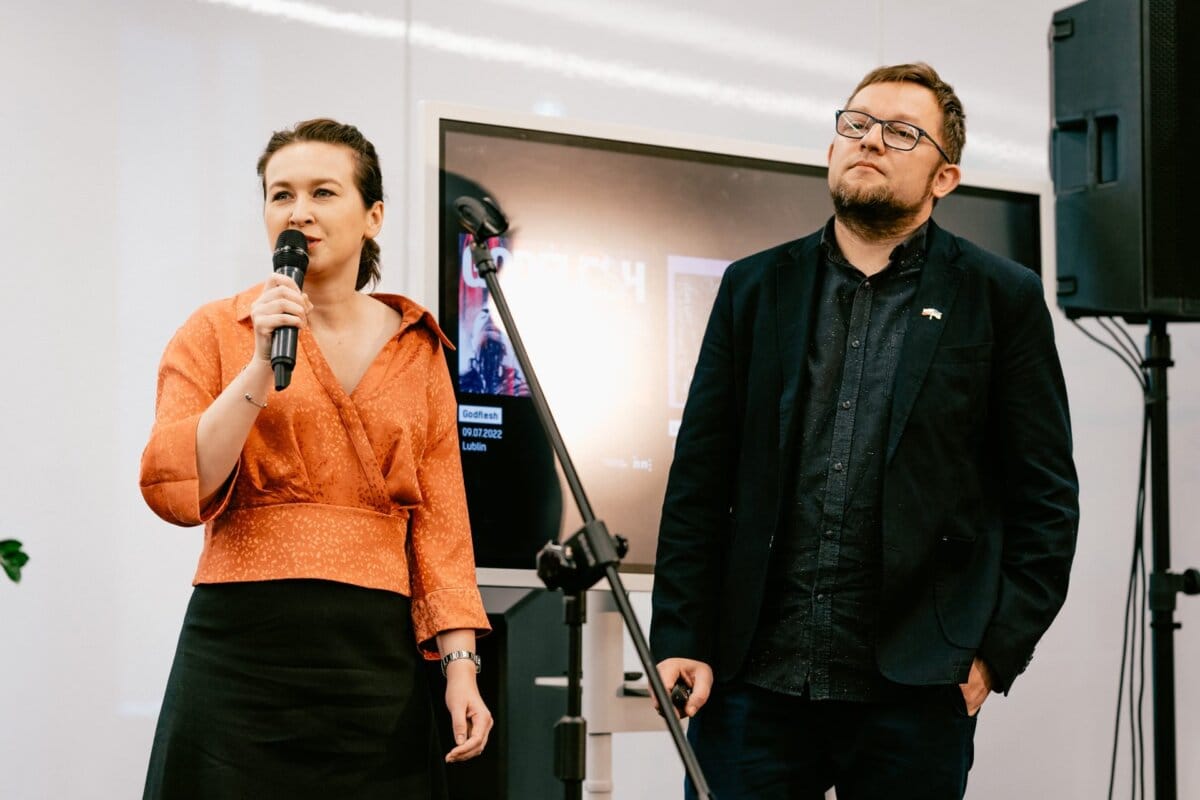 A woman dressed in a red blouse and black skirt holds a microphone and speaks, while Marcin Krokowski, a well-known photographer in Warsaw, stands next to her. Behind them is a screen displaying a presentation in a modern room with white walls, capturing the essence of this photographed event moment. 