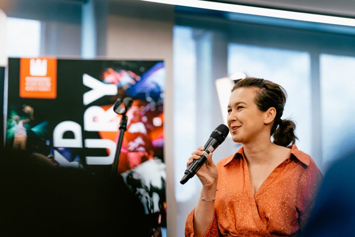 A person with long hair tied back, wearing an orange blouse, speaks into a microphone at an event taking place in a room. A colorful banner with text and images is visible in the background. The room appears well lit thanks to the large windows, which was beautifully captured by Marcin Krokowski, a photographer in Warsaw.  