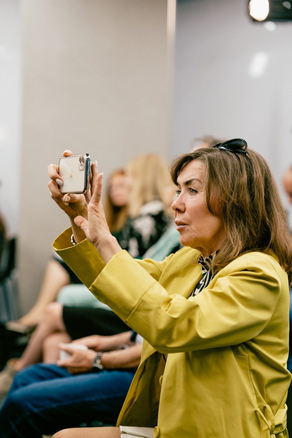 A woman wearing a yellow jacket and sunglasses on her head takes a photo with a smartphone. She is sitting among other out-of-focus people in the background, suggesting a public gathering or event, likely captured by well-known Warsaw photographer Marcin Krokowski. 