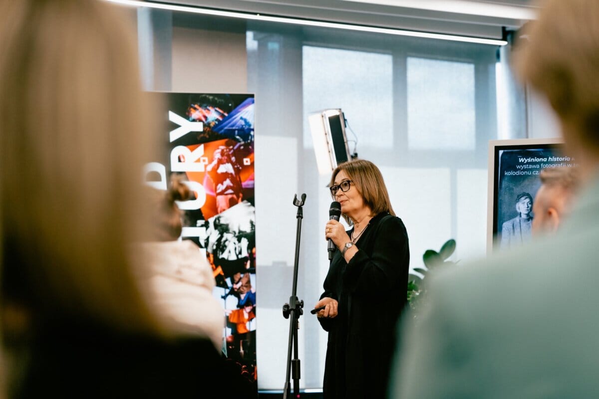 A woman with shoulder-length hair stands and speaks into a microphone in a well-lit room. Behind her, a colorful vertical banner reading "HISTORY" is partially visible. A blurred viewer's arm appears in the foreground, capturing the essence of Marcin Krokowski's eventful moment in Warsaw.  