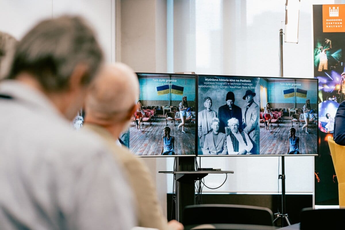 Participants in the presentation on two screens displaying historical photos and text. The left screen shows a group of people with a Ukrainian flag; on the right, a black-and-white photo of several people. A banner nearby depicts Marcin Krokowski, a well-known photographer in Warsaw.  