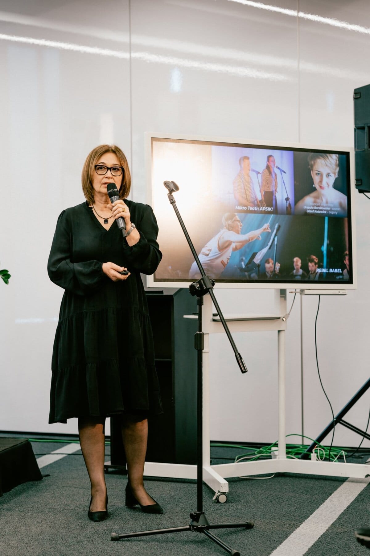 A woman in a black dress stands in front of a microphone and speaks to an engaged audience. Behind her, images of various speakers and performers are projected on a screen. She is in a modern conference room with white walls and a gray carpet, meticulously captured by Marcin Krokowski, an event photographer.  