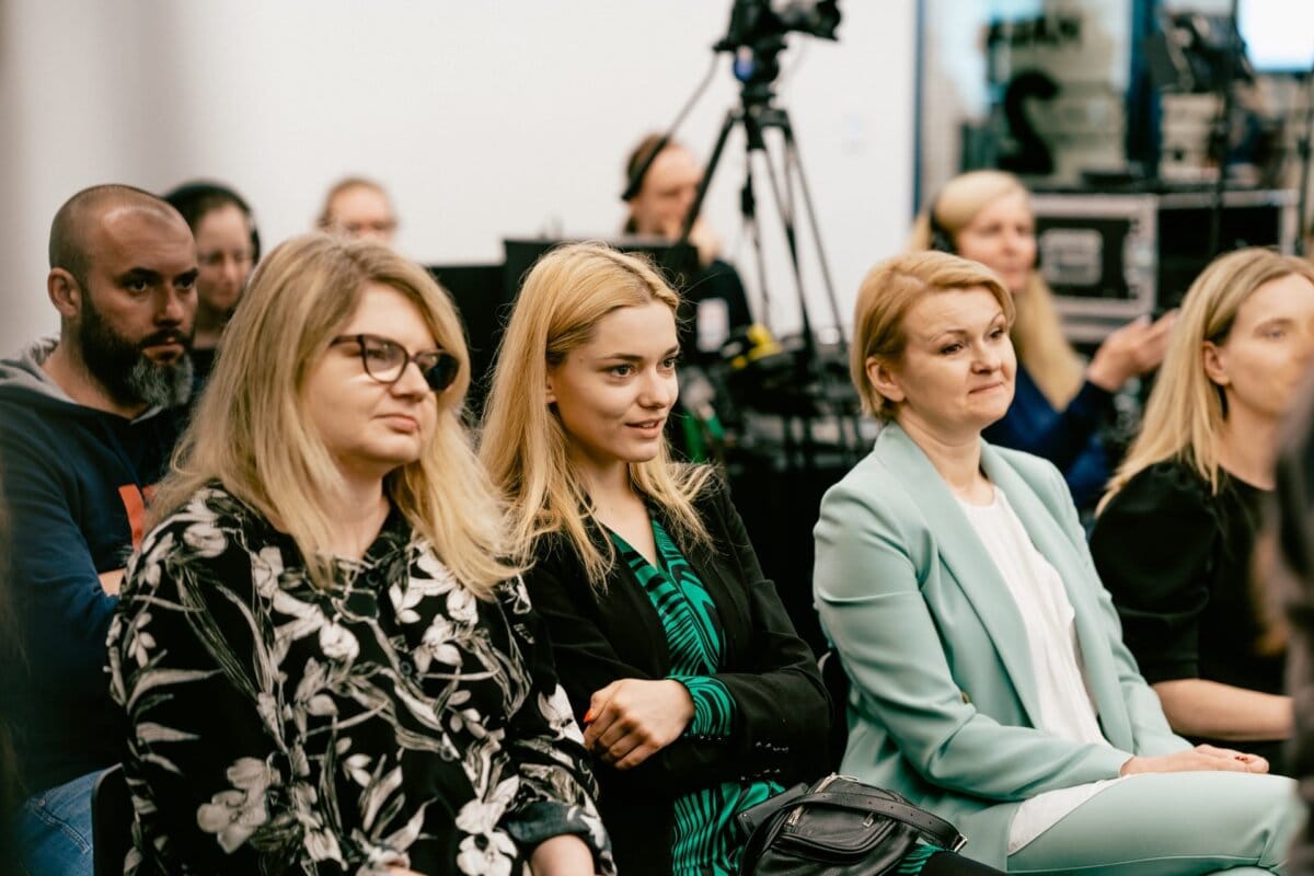 A group of people are sitting attentively in a closed room. In the front row sit three women, two with blond hair and one with glasses. Behind them, others can be seen, including a bearded man and a crew member operating a camera. The atmosphere seems formal, as captured perfectly by Marcin Krokowski, a photographer in Warsaw.   
