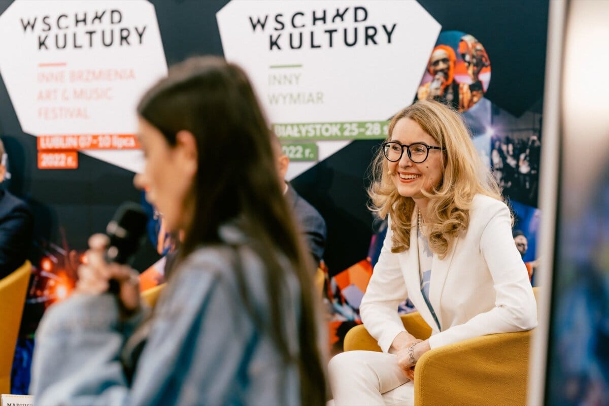 A woman in a white suit and glasses sits on a chair and smiles, while in the foreground a blurred figure speaks into a microphone. In the background are banners with Polish text promoting an art and music festival, captured by event photographer Marcin Krokowski. 