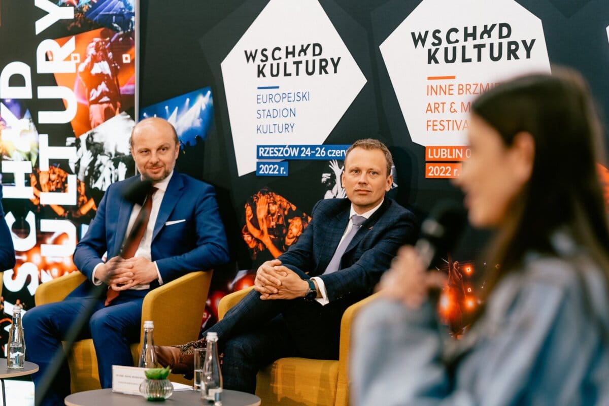 During the panel discussion, two men in suits sit on the stage and a woman speaks into the microphone, somewhat blurred in the foreground. Behind them, banners display text about the "East of Culture" festival with details of the event indicated for Rzeszow and Lublin in 2022. (Photo: Marcin Krokowski)  