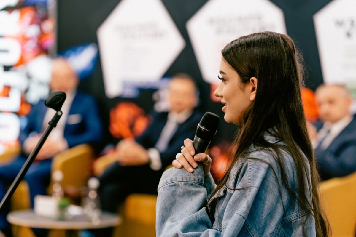 A woman with long dark hair speaks into a microphone at the event. She is wearing a light denim jacket. In the background, several men in suits sit on stage, with smudged banners behind them. The atmosphere suggests a formal or professional setting captured by Marcin Krokowski, a photographer in Warsaw.   