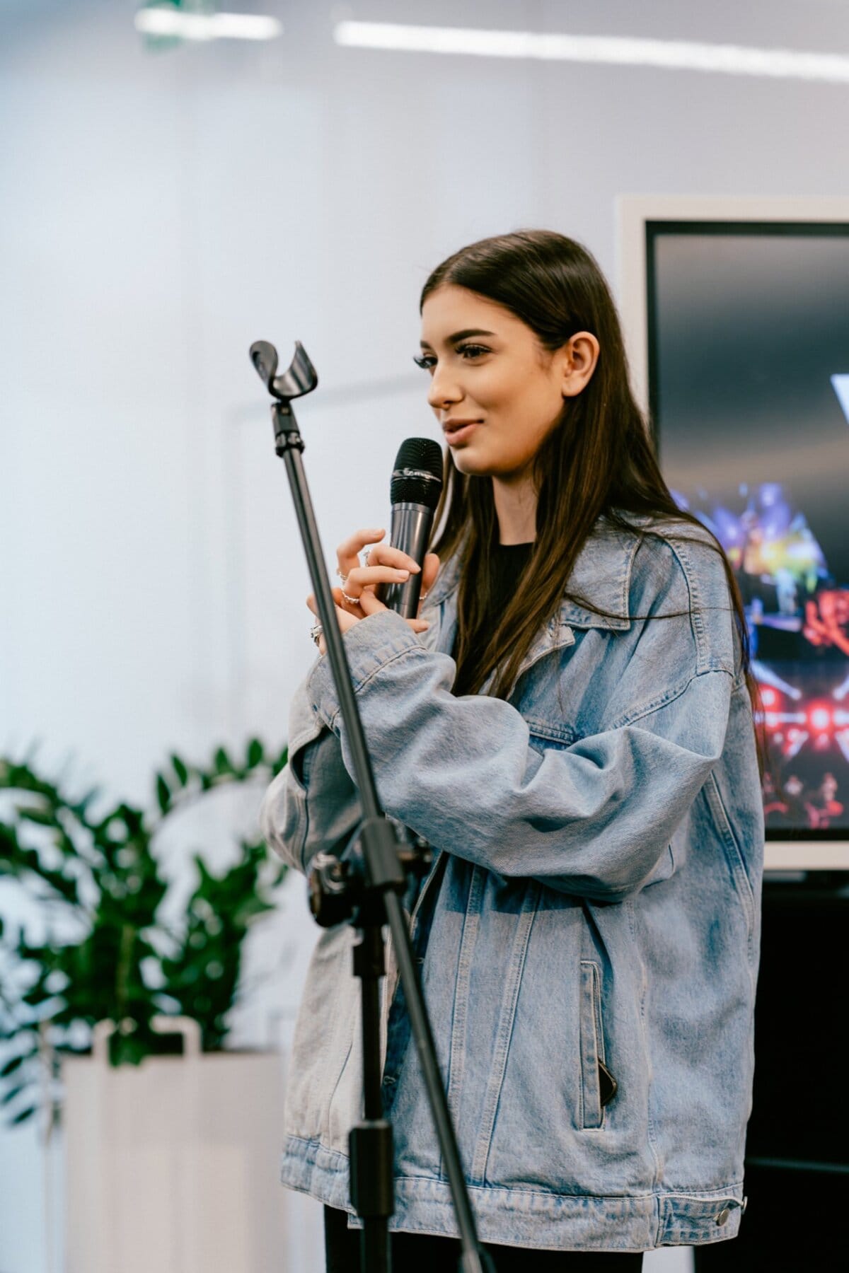 A young woman with long dark hair speaks into a microphone while standing in front of a large screen. She is wearing a light blue denim jacket and standing next to a black microphone stand. In the background are green plants and a white wall, perfectly captured by Marcin Krokowski, an event photographer in Warsaw.  