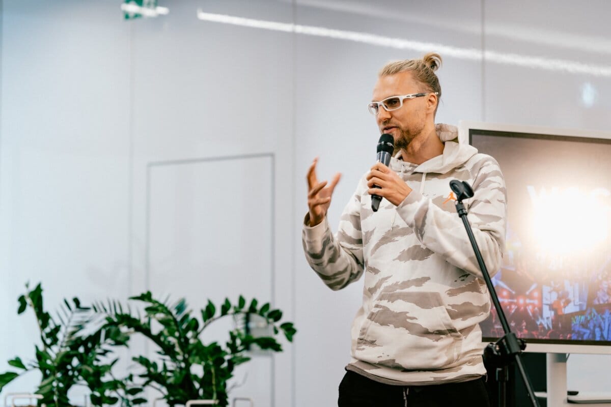 Marcin Krokowski, a well-known event photographer, wearing a patterned hoodie and glasses, speaks into a microphone and gestures with his hand. He stands in a modern, well-lit room with a large screen in the background and green plants on the sides. 