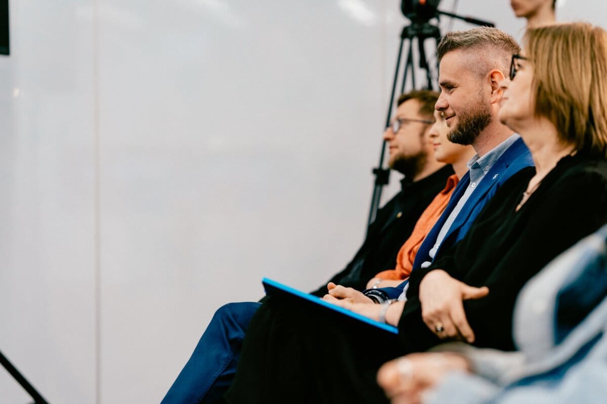 A row of seated people carefully watching an event or presentation. A man in a blue suit and a woman in glasses are in focus, others are slightly blurred. A camera on a tripod is visible in the background, capturing moments with the precision characteristic of prominent Warsaw photographer Marcin Krokowski.  