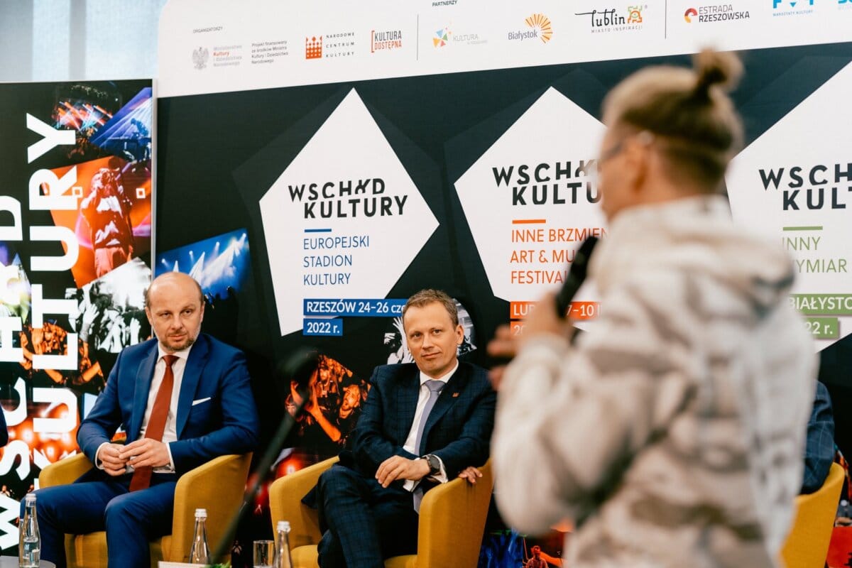 Three people sit on stage at the East of Culture event. Two men in suits sit on chairs and listen to the person in the foreground speak into a microphone. Banners in the background display event information captured by a prominent photographer in Warsaw, Marcin Krokowski. The atmosphere is formal and professional.   