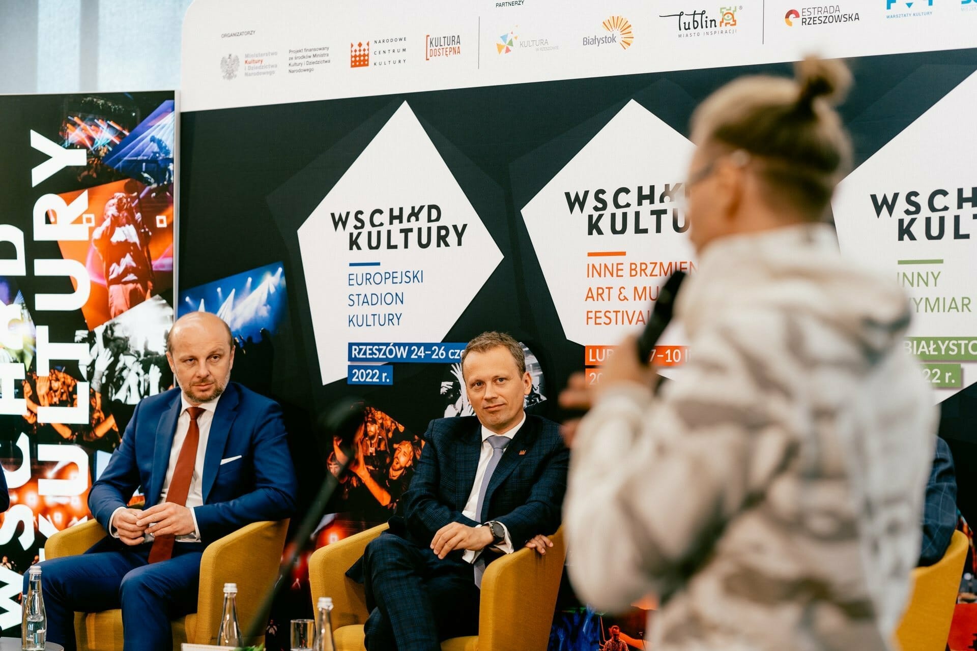 Biało-pomarańczowy samochód sportowy jest wystawiony w nowoczesnym salonie podczas otwarcia salonu, z dużymi oknami, szarymi płytkami podłogowymi i kilkoma osobami w tle, niektórzy robią zdjęcia.