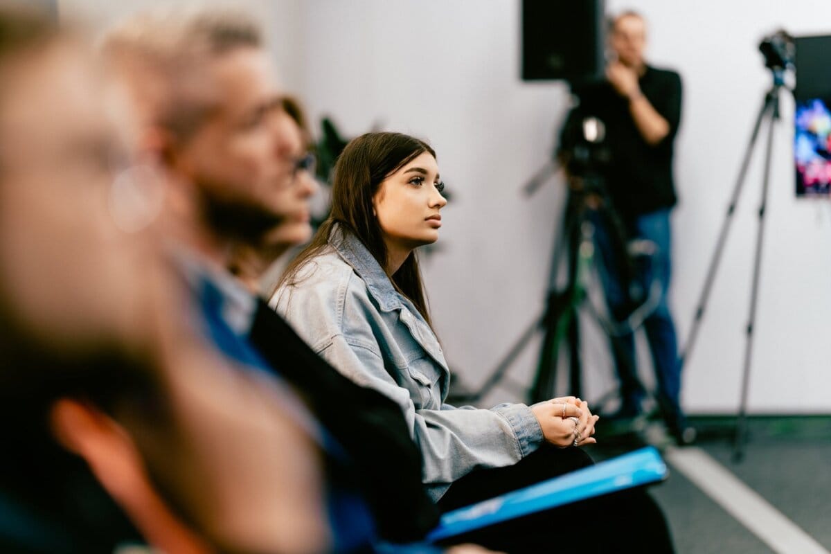 A young woman with long, dark hair listens intently during the event. She sits in the crowd, focused, wearing a gray jacket and holding a notebook. Cameras and other participants can be seen in the background; it seems to have been captured by Marcin Krokowski, a well-known event photographer in Warsaw.  