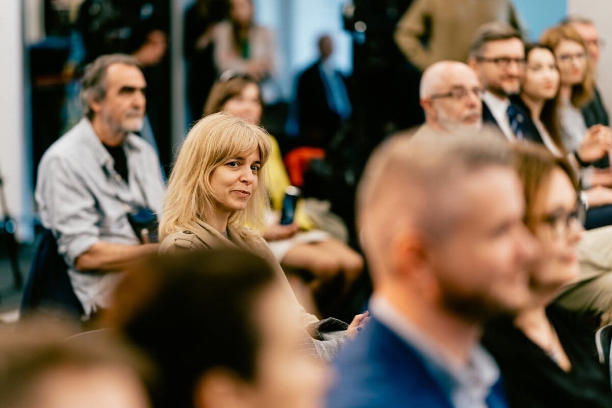 During what looks like a conference or seminar, a group of people are sitting and listening intently. A smiling woman with blond hair stands out in the foreground. In the background are men and women with notepads and electronic devices, ingeniously captured by well-known Warsaw photographer Marcin Krokowski.  