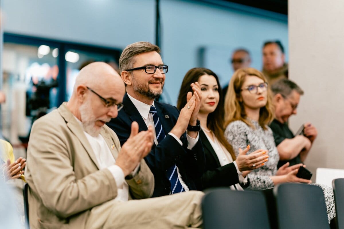 A group of people sitting in a well-lit room and applauding. In the foreground is a smiling man wearing a suit and clapping his glasses. Also clapping next to him are a man in a beige suit and a woman with dark hair. Visible in the background are the other participants, beautifully captured by event photographer Marcin Krokowski.   