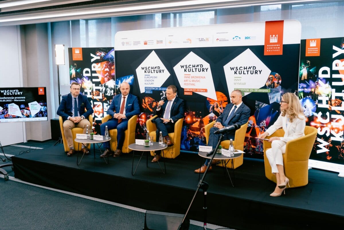   Five people sit on stage during a panel discussion at a cultural festival. They sit in mustard-colored chairs against a backdrop of the event's logo and vivid design. Four of them are in suits and one is wearing a white outfit. Between them on the table are microphones and water bottles, which was perfectly captured by Marcin Krokowski, a respected photographer in Warsaw who specializes in   