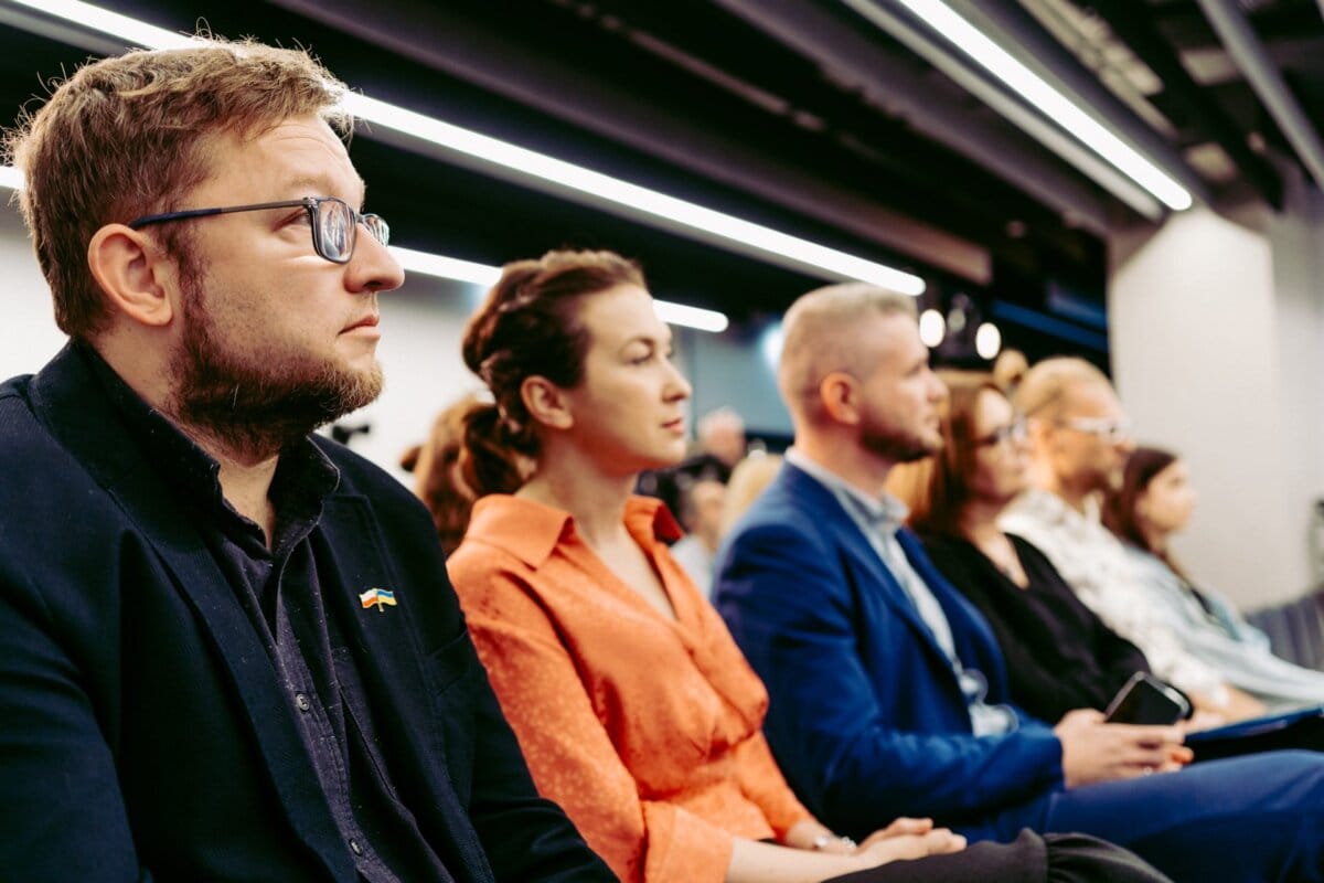 A group of people are sitting in a conference room and listening intently. The focus is on four people in the front row, two men and two women, dressed in formal attire. Captured by Marcin Krokowski, a photographer in Warsaw, the room is well lit with overhead lamps, creating a professional atmosphere.  