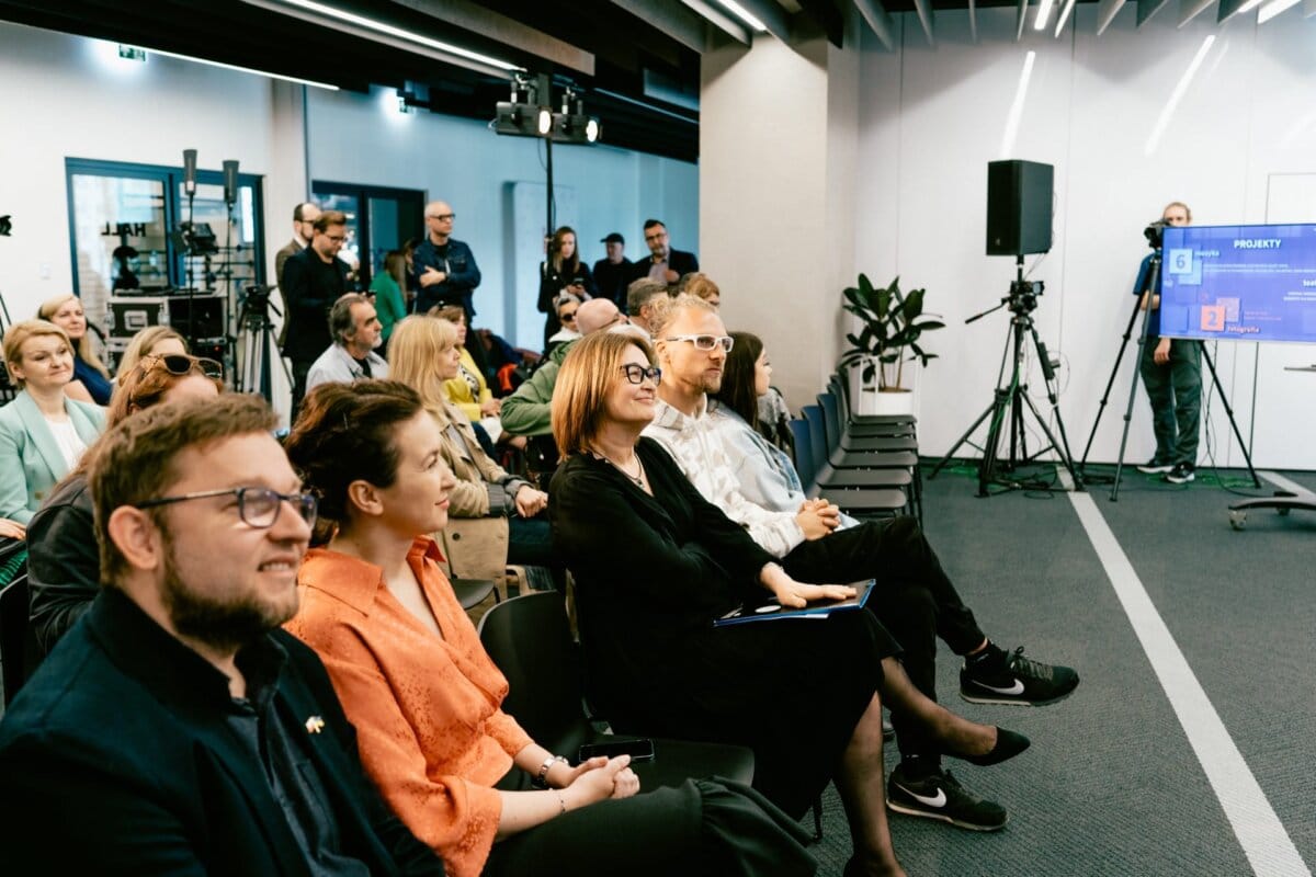 A diverse audience, seated and attentive, watches the presentation in a modern conference room. In the background, the speaker stands in front of a screen with visuals, while Marcin Krokowski, a photographer in Warsaw, along with other photographers and videographers capture the event. The atmosphere is professional and committed.  