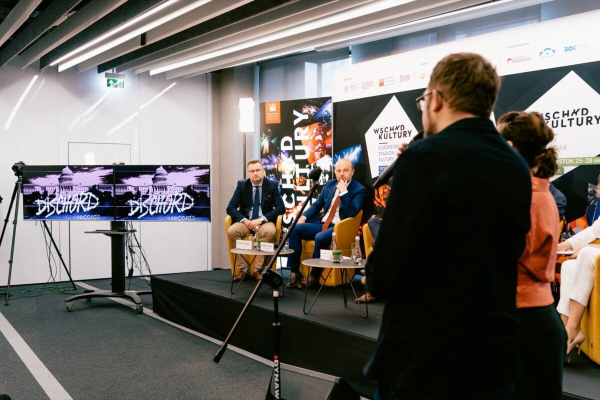 A panel of five speakers sits on stage, including event photographer Marcin Krokowski, who addresses the audience during the conference. A large screen next to them displays graphics, while the audience stands and asks questions. The setting is a modern conference room with a projector and speakers.  