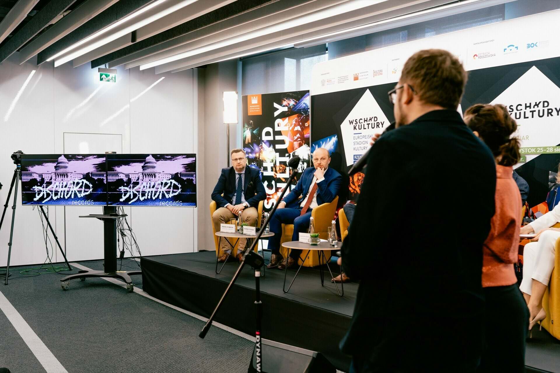 Fünf Podiumsteilnehmer sitzen auf der Bühne, darunter Eventfotograf Marcin Krokowski, der während einer Konferenz zum Publikum spricht. Neben ihnen wird eine Grafik auf der großen Leinwand gezeigt, und Teilnehmer stehen auf und stellen Fragen. Der Rahmen ist ein moderner Konferenzraum mit Projektor und Lautsprechern.