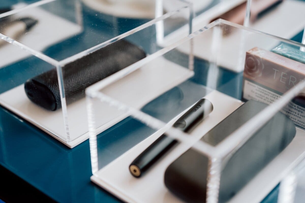 Close-up of several electronic devices and accessories housed in clear acrylic boxes, as if captured by a photographer at conferences. Elements include a sleek black cylindrical device, a black rectangular case and a small box with visible text. The display is set on a glossy blue surface.  