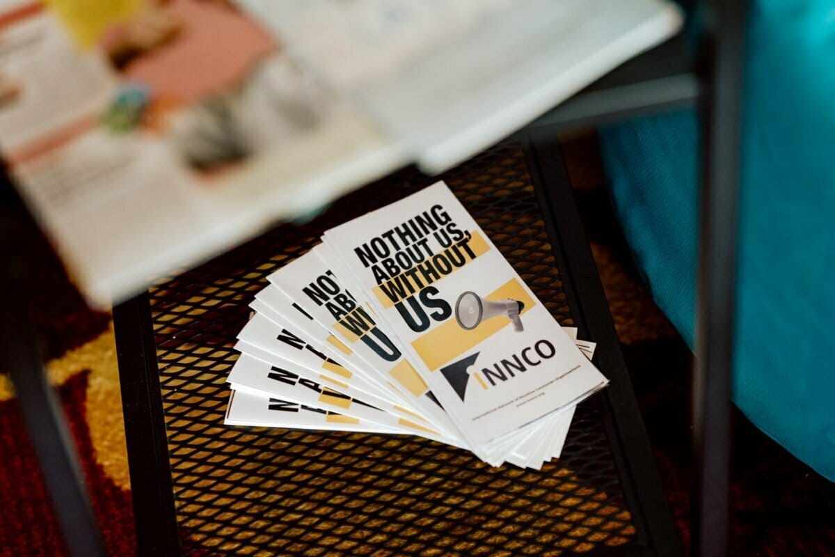 A stack of brochures on a black metal table with the text "NOTHING ABOUT US WITHOUT US" written in bold black and gold letters. The brochures also contain a gray illustration of a hearing aid and the "ANNCO" logo. A blurry magazine, perhaps captured by a photographer at the conferences, is partially visible in the background.  