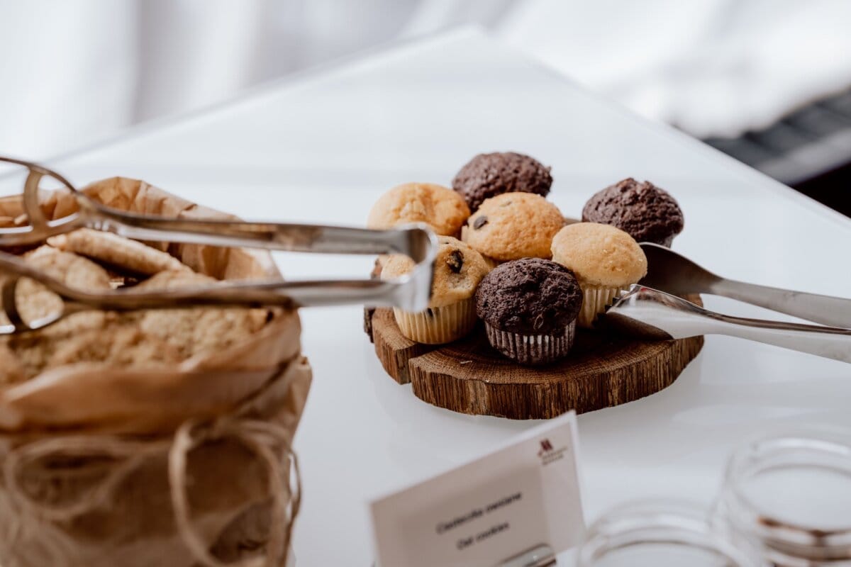 A plate of assorted mini cupcakes, including chocolate and vanilla flavors, is displayed on a wooden surface. Metal tongs rest on the plate. On the left, a brown paper bag containing the cookies is partially visible. The background is blurred for a soft-focus effect, ideal for photographers at conferences looking for stunning detail shots.   